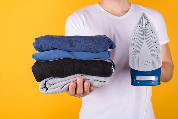 Concepto de planchado de ropa. Foto recortada de un hombre sosteniendo una pila de ropa doblada y una plancha aislada sobre fondo amarillo