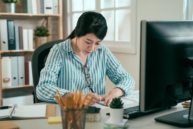 concepto de planificación de trabajo de diseño de ingeniera arquitecta asiática. joven china sentada en el escritorio en compañía contra la ventana interior. elegante trabajador interior usando regla y dibujo de pluma en la computadora