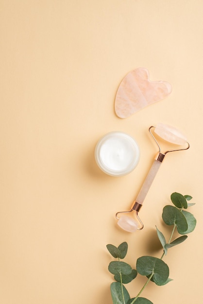 Concepto de procedimiento de belleza Foto vertical de vista superior de rodillo de cuarzo rosa masajeador gua sha tarro de crema de vidrio y rama de eucalipto sobre fondo beige pastel aislado