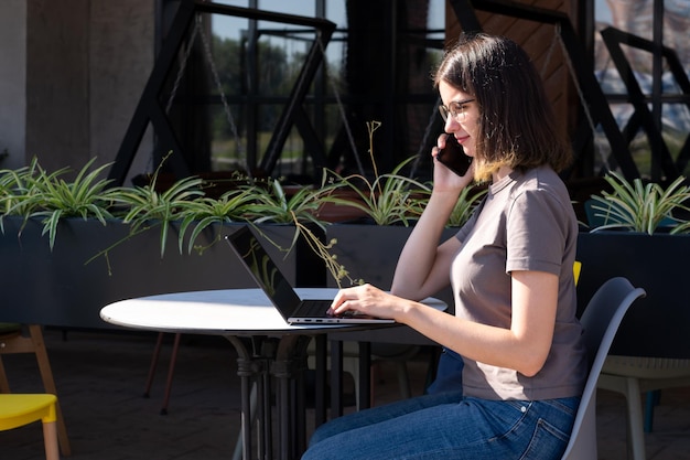 El concepto de un profesional independiente Una hermosa estudiante morena con anteojos está hablando por teléfono usando una computadora portátil en un café