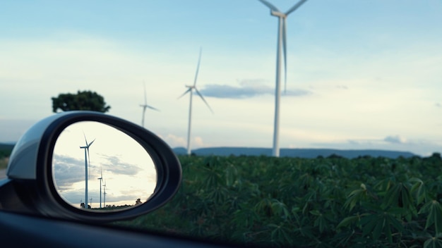 Concepto progresivo de infraestructura energética futura de turbina eólica reflejado en el espejo lateral de un vehículo eléctrico que se carga en una estación de carga alimentada por energía verde y renovable de una turbina eólica