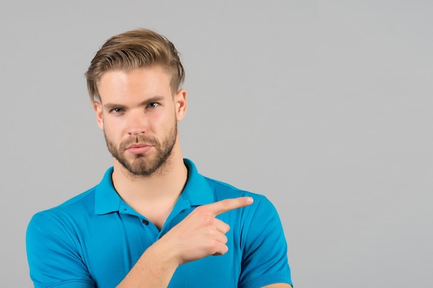 Concepto de publicidad de asesoramiento. Guy barbudo atractivo dedo índice apuntando a un lado. El rostro estricto del hombre se ve con confianza, fondo gris. Hombre con barba sin afeitar se ve guapo bien arreglado.