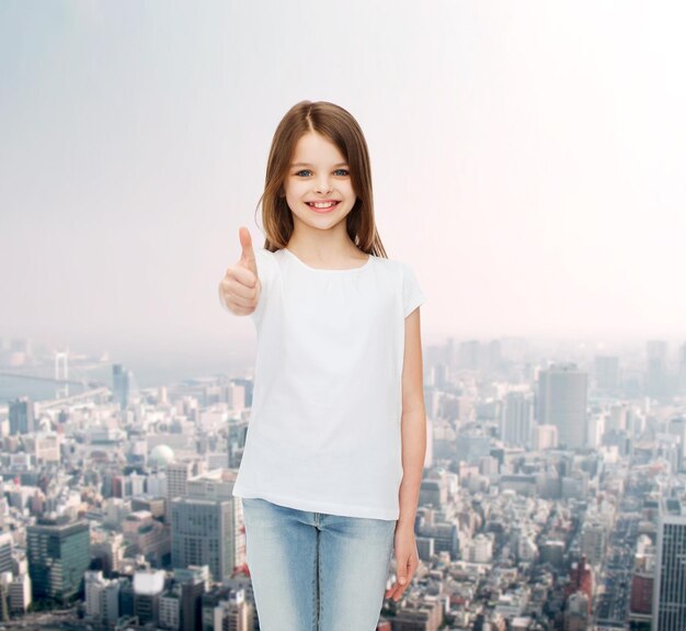 concepto de publicidad, infancia, gestos y personas - niñita sonriente con camiseta blanca mostrando los pulgares hacia arriba sobre el fondo de la ciudad