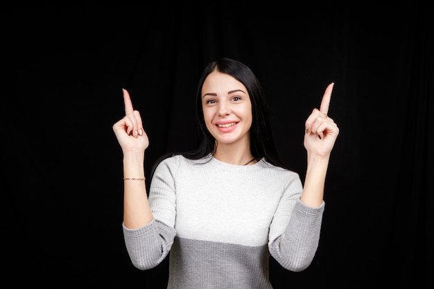 El concepto de publicidad. Una mujer positiva, bonita y casual con una sonrisa radiante y un suéter brillante en un espacio negro indica dos dedos índices y mira el espacio vacío de la copia.