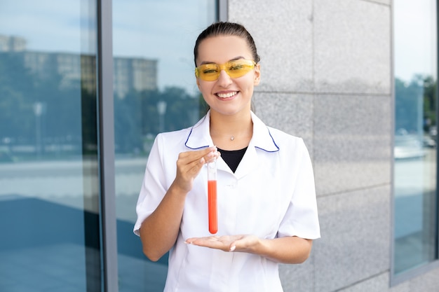 Concepto de química, ciencia, medicina y biología - Química femenina blanca europea con una bata blanca y gafas amarillas sosteniendo un tubo de ensayo de líquido rojo y realizando una investigación al aire libre en un clima soleado