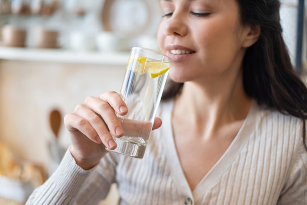 Concepto de refresco joven bebiendo agua con limón sosteniendo un vaso de pie en la cocina closeup