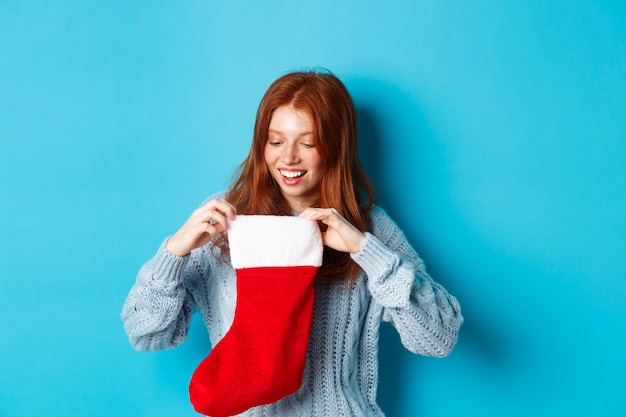 Concepto de regalos y vacaciones de invierno. Chica pelirroja divertida mirando dentro de la media de Navidad y sonriendo feliz, recibiendo regalo de Navidad, de pie contra el fondo azul.