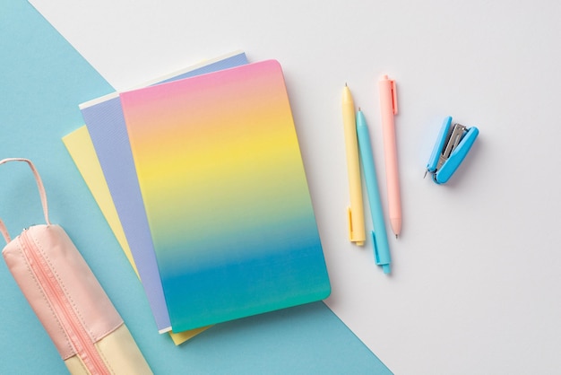 Concepto de regreso a la escuela Foto de vista superior de útiles escolares bolígrafos de cuaderno de color degradado mini grapadora y estuche rosa sobre fondo bicolor azul y blanco