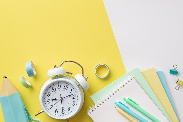 Concepto de regreso a la escuela Foto de vista superior de útiles escolares reloj despertador cuadernos coloridos estuche bolígrafos clips de carpeta y cinta adhesiva sobre fondo bicolor amarillo y blanco