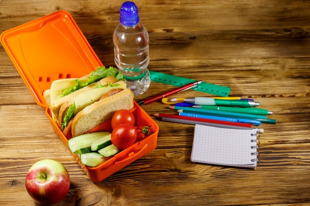 Concepto de regreso a la escuela Útiles escolares botella de agua manzana y lonchera con sándwiches y verduras frescas en un escritorio de madera