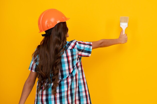 Foto concepto de renovación y construcción de niños niña pequeña con casco protector y pincel para pintar paredes aisladas en fondo amarillo