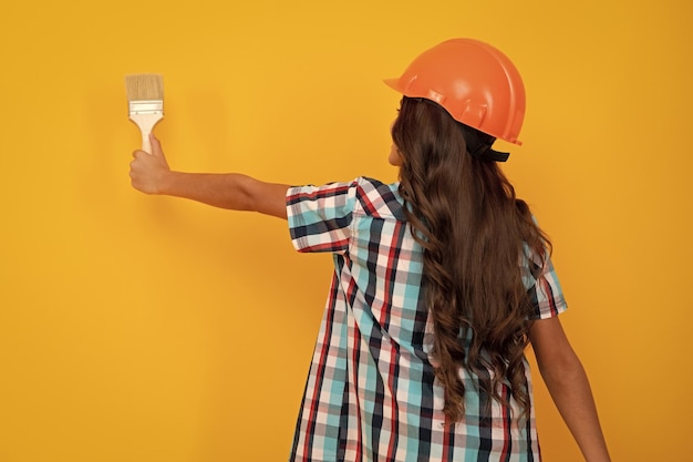 Concepto de renovación y construcción de niños Niña pequeña con casco protector y pincel para pintar paredes aisladas en fondo amarillo