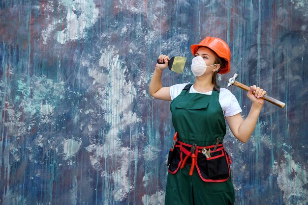 Concepto de renovación y remodelación. Mujer con casco y máscara protectora posando con martillo y espátula