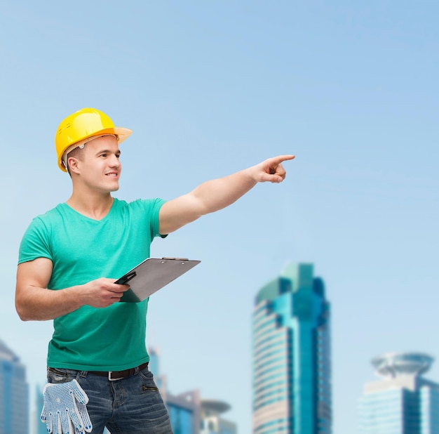 concepto de reparación, construcción y mantenimiento - hombre sonriente en casco con portapapeles apuntando con el dedo