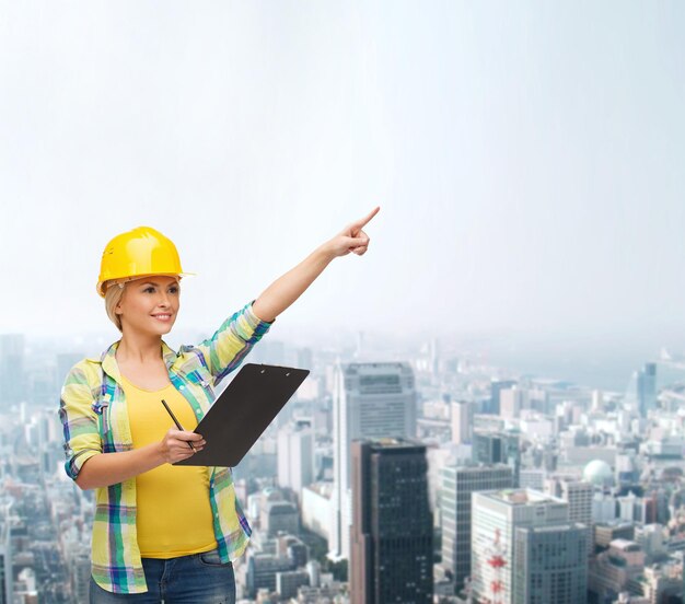 concepto de reparación, construcción y mantenimiento - mujer sonriente en casco con portapapeles apuntando con el dedo