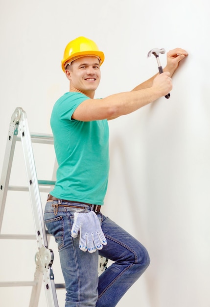 concepto de reparación, construcción y renovación del hogar - hombre sonriente con casco protector amarillo martillando clavos en la pared