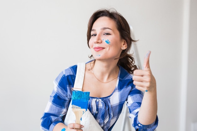 Concepto de reparación, renovación, trabajador y personas - pared de pintura de mujer joven feliz en casa, mostrándonos los pulgares para arriba.