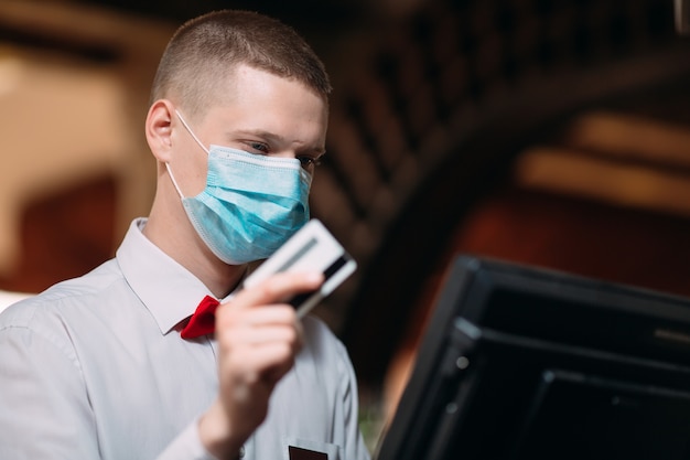 Concepto de restaurante, personas y servicio. Hombre o camarero en máscara médica en mostrador con caja trabajando en el bar o cafetería.