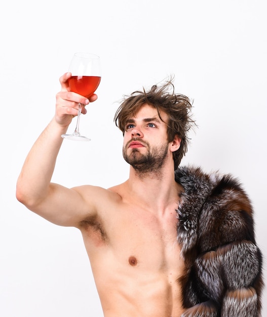 Concepto de riqueza y lujo. Chico atractivo posando abrigo de piel sobre el cuerpo desnudo. Vino de bebida de pelo despeinado macho soñoliento sexy o alcohol aislado en blanco. Símbolo de estatus de lujo. Estilo de vida de lujo y bienestar.