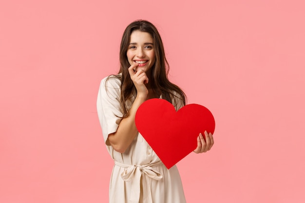 Concepto de romance, belleza y felicidad. Sensual seductora, coqueta joven vestida, tocando los labios, expresa deseo o tentación, sonriendo quiere probar algo, sosteniendo un gran corazón rojo, el día de San Valentín