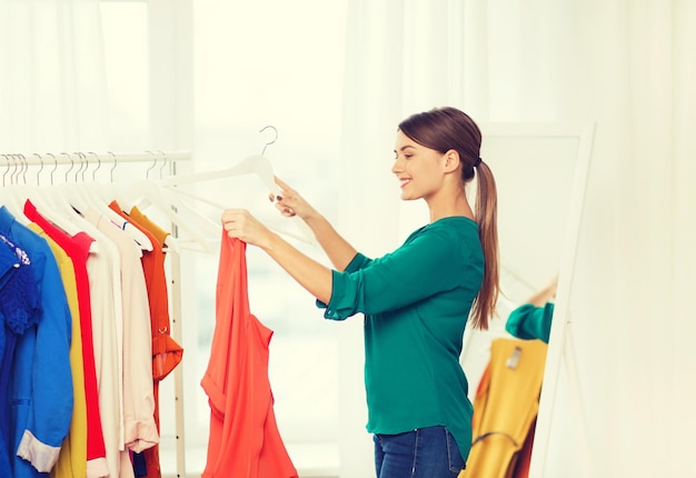 Concepto de ropa, moda, estilo y personas - mujer feliz eligiendo ropa en el armario de casa