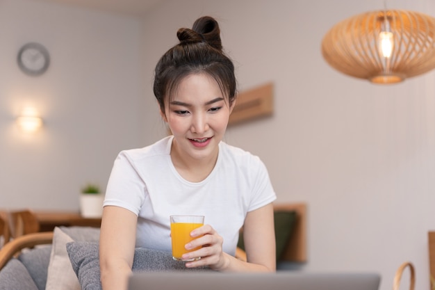 Concepto de sala de estar en la cafetería una chica con un moño usando su computadora portátil en Internet con un vaso de jugo de naranja.