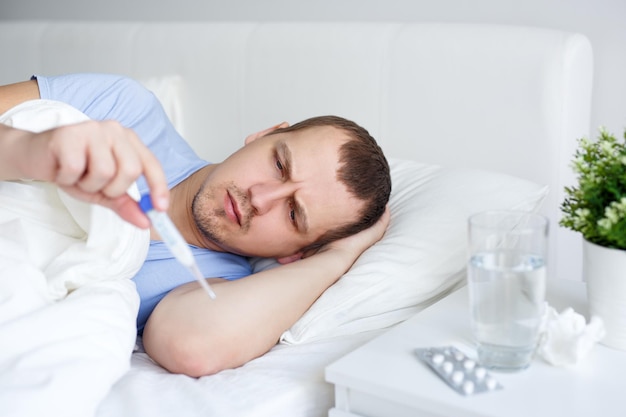 Concepto de salud y enfermedad hombre enfermo con fiebre acostado en la cama y mirando el termómetro