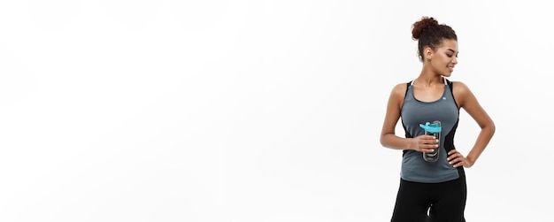 Concepto de salud y fitness hermosa chica afroamericana en ropa deportiva sosteniendo una botella de agua después del entrenamiento aislado sobre fondo blanco de estudio