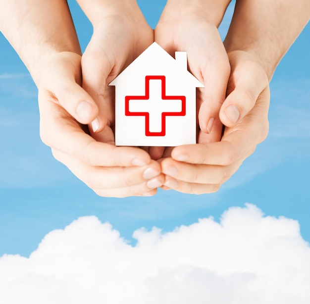 concepto de salud, medicina y caridad - manos masculinas y femeninas sosteniendo una casa de papel blanco con un signo de cruz roja