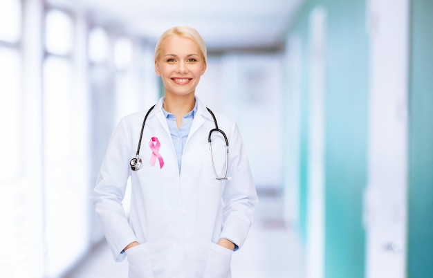 concepto de salud y medicina - doctora sonriente con estetoscopio y cinta rosa de concientización sobre el cáncer sobre los antecedentes del hospital