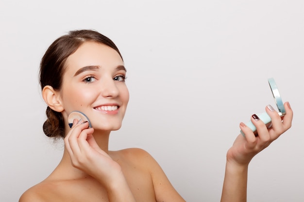 Concepto de salud, personas y belleza: mujer joven que aplica colorete en la cara con borla de polvo, concepto de cuidado de la piel, composición fotográfica de una niña morena, aislada sobre fondo blanco