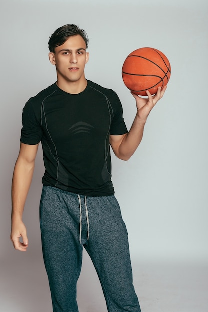 Concepto de salud, personas, deporte y estilo de vida: jugador real. Hombre sonriente joven hermoso que lleva una pelota de baloncesto y que mira la cámara mientras está de pie contra el fondo gris