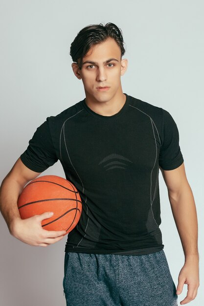 Concepto de salud, personas, deporte y estilo de vida: jugador real. Hombre sonriente joven hermoso que lleva una pelota de baloncesto y que mira la cámara mientras está de pie contra el fondo gris