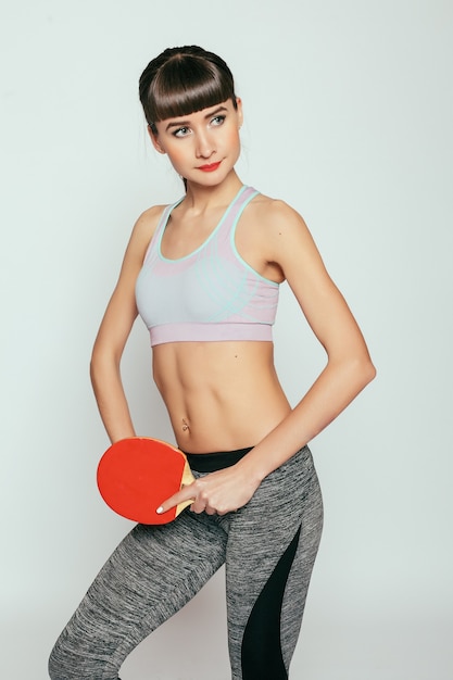 Concepto de salud, personas, deporte y estilo de vida - mujer joven sosteniendo una raqueta de tenis de mesa y una pelota. Chica joven fitness feliz con cuerpo deportivo en el estudio sobre un fondo gris.