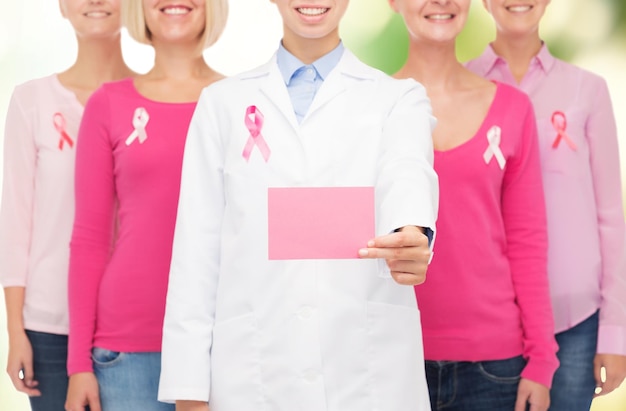 Foto concepto de salud, personas y medicina - cierre de mujeres sonrientes en camisas con cintas rosas de concientización sobre el cáncer de mama y tarjeta de papel en blanco sobre fondo verde