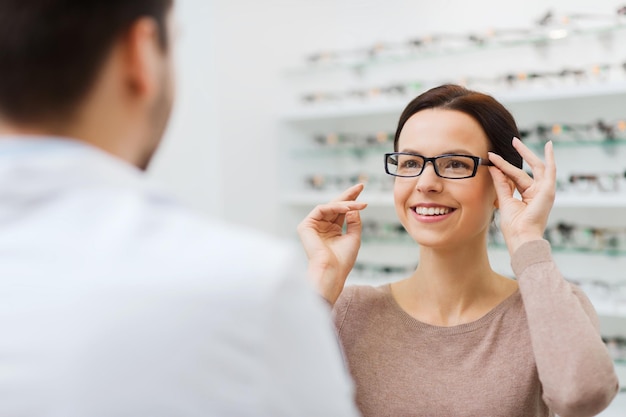 concepto de salud, personas, vista y visión - mujer feliz con óptico eligiendo anteojos en la tienda de óptica