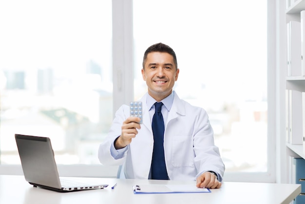 concepto de salud, profesión, personas y medicina - médico sonriente con bata blanca con tabletas y computadora portátil en el consultorio médico