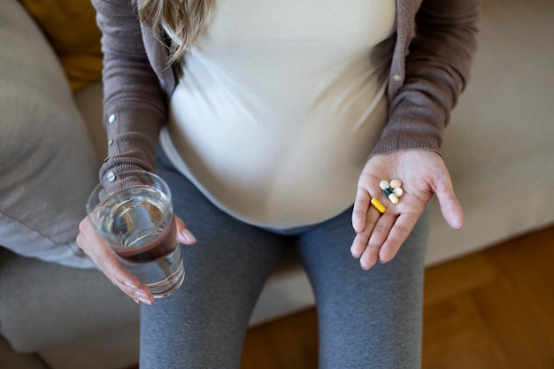 Concepto de salud. Vista recortada de la mujer embarazada tomando píldoras de vitamina, sosteniendo la taza con agua en las manos, sentada sola detrás de la mesa de madera