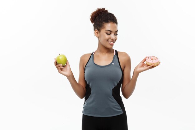 Concepto saludable y de dieta - hermosa afroamericana deportiva toma una decisión entre rosquilla y manzana verde aislada en una persona de fondo blanco