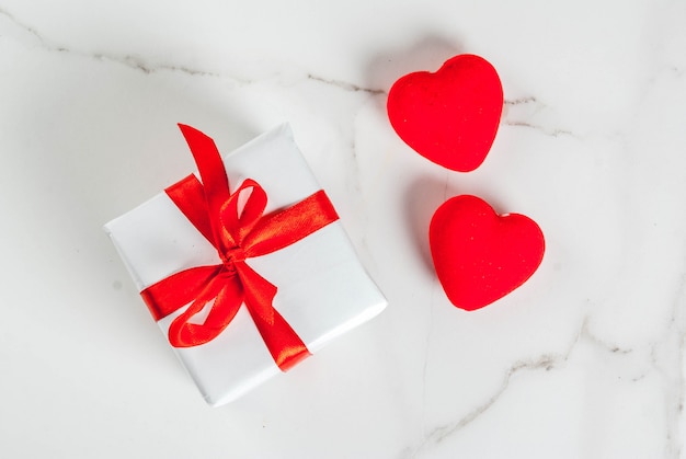 Concepto de San Valentín, caja de regalo envuelta en blanco con cinta roja y corazones de terciopelo rojo, sobre fondo de mármol blanco, vista superior del espacio de copia