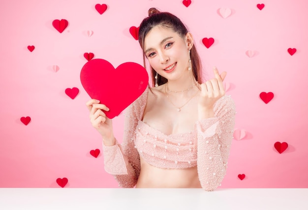 Concepto de San Valentín Mujer sexy asiática joven reunida en cola de caballo con maquillaje natural en la cara tiene labios regordetes y piel limpia y fresca que muestra el signo del amor en el fondo del patrón de corazón rosa