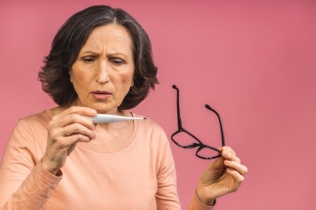 Concepto de síntoma de coronavirus. Mujer de edad avanzada madura con gripe sosteniendo un termómetro electrónico en la mano, que se encuentran aisladas sobre fondo rosa con espacio de copia