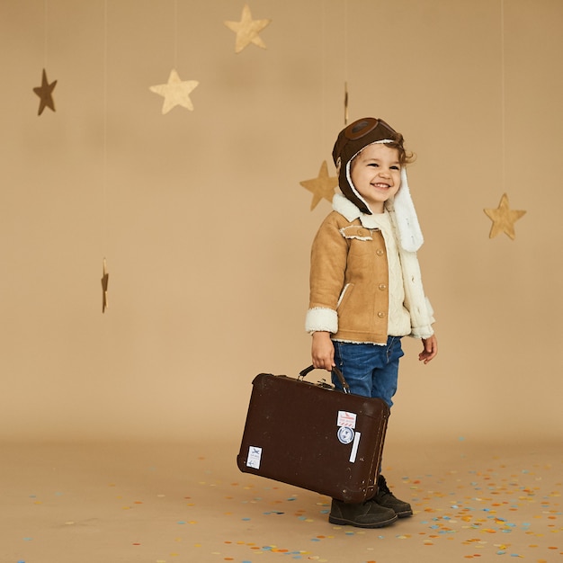 Concepto de sueños y viajes. Niño piloto aviador con un avión de juguete y una maleta juega en una superficie beige