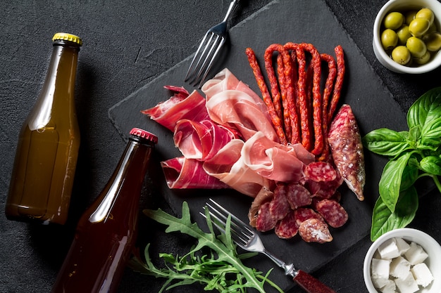 Foto concepto de tapas. jamón, chorizo, cerveza embotellada, aceitunas, rúcula, albahaca y queso sobre una pizarra negra. concepto antipasti. copia espacio