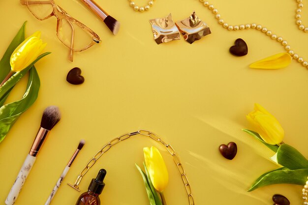 Concepto de tarjeta de belleza Pinceles de maquillaje de moda gafas suero cuentagotas cadena de perlas y aretes Accesorios dorados sobre fondo amarillo brillante con corazones de chocolate y tulipán amarillo Copiar espacio