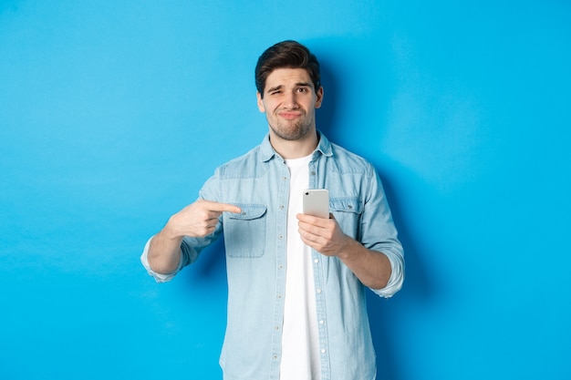 Concepto de tecnología, aplicaciones y compras online. Chico escéptico y disgustado apuntando con el dedo al teléfono inteligente y haciendo muecas decepcionado, de pie sobre fondo azul.