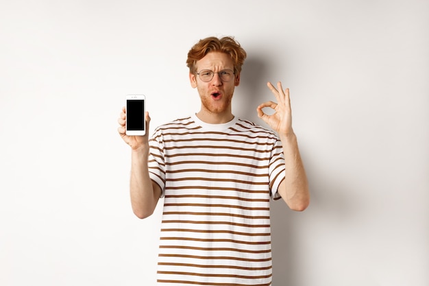 Concepto de tecnología y comercio electrónico. Hombre joven con el pelo rojo que muestra el signo bien y la pantalla del teléfono inteligente en blanco, alabando una aplicación impresionante o un videojuego, fondo blanco