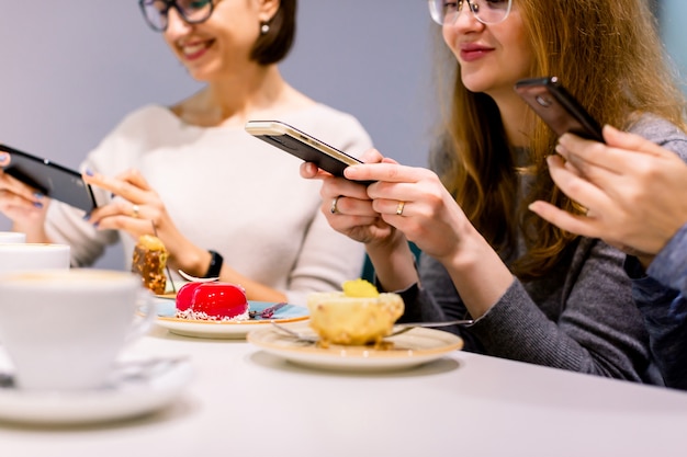 Concepto de tecnología, estilo de vida, amistad y personas: tres mujeres jóvenes felices con teléfonos inteligentes haciendo fotos de sus tazas de café y postres en un café en el interior