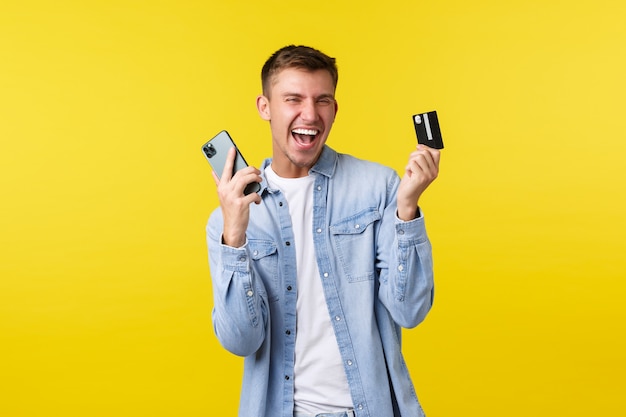 Concepto de tecnología, estilo de vida y publicidad. Hombre guapo alegre feliz bailando como compra increíble en línea, mostrando tarjeta de crédito y teléfono móvil como regocijo, fondo amarillo.