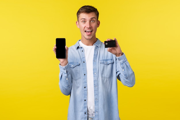 Concepto de tecnología, estilo de vida y publicidad. Hombre guapo emocionado y optimista que muestra la tarjeta de crédito y la pantalla del teléfono inteligente, presenta una nueva aplicación o tienda en línea, recomendándola, fondo amarillo.